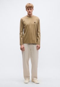 Hosszú ujjú, világosbarna ing kerek nyakkivágással és apró fekete logóval, bézs, bőszárú nadrággal és krémszínű tornacipőkkel párosítva.