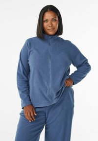 Sininen fleece-takki, jossa on täysi vetoketju ja korkea kaulus. Pehmeä rakenne, pitkät hihat ja rento istuvuus. Yhdistetty matchaaviin housuihin.