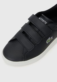 Černé kožené tenisky s třemi suchými zipy, bílou gumovou podrážkou a logem Lacoste znázorněným jako zelený krokodýl na boku.
