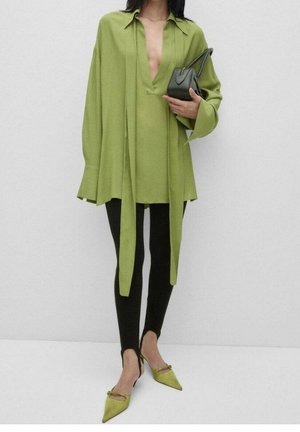 Person iført en løs limegrøn tunika med lange snører, sorte tætsiddende bukser, limegrønne spidse sko og holder en lille mørkegrøn håndtaske.