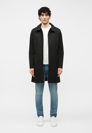 Fiatal férfi göndör hajjal, fekete kabátot, fehér pólót, kék farmert és fehér tornacipőt viselve, egy egyszínű világos háttér előtt áll.