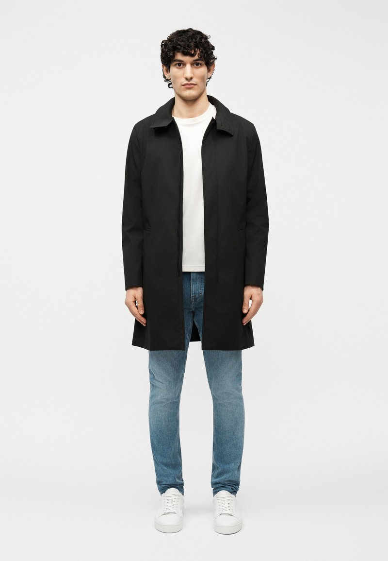 Νέος άνδρας με σγουρά μαλλιά, ντυμένος με μαύρο παλτό, λευκό T-shirt, μπλε τζιν και λευκά αθλητικά παπούτσια, στέκεται μπροστά σε ένα απλό φωτεινό φόντο.