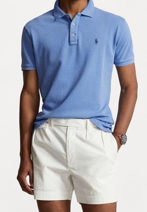 Homme portant un polo bleu clair à manches courtes avec col et un short plissé blanc, montre-bracelet au poignet gauche, main droite dans la poche.