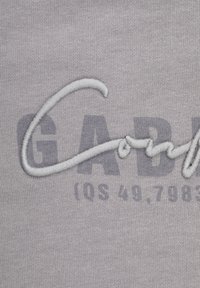 Grått tyg med en broderad "GAD"-logotyp. Designen har en strukturerad, upphöjd söm och en skuggbeskrivning nedanför.
