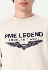Bež dukserica s tamnoplavim grafičkim motivom koji sadrži natpis "PME LEGEND AMERICAN CLASSIC" i dizajn krilate štit. Glatka tkanina i okrugli izrez.
