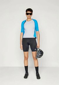 Persona in abbigliamento da ciclismo con pantaloni corti neri, maglia grigia, giacca blu, scarpe da ciclismo nere e occhiali da sole, che tiene un casco da bici grigio.