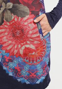 Blommönstrad klänning i röda, blå och gröna nyanser, med en ficka och varierade texturer från släta till något upphöjda mönster.