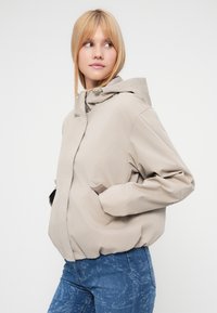 Μπεζ μπουφάν με κουκούλα, με λεία υφή, άνετη εφαρμογή και πλαϊνές τσέπες. Φορεμένο πάνω από μπλε τζιν με μοτίβο, υπογραμμίζοντας το στιλ με στρώσεις ρούχων.