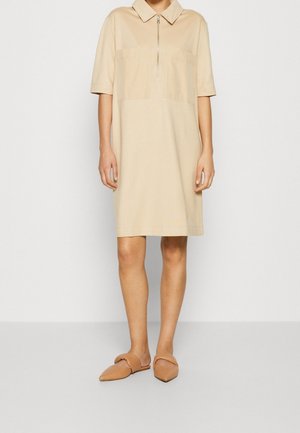 Person iført en beige kortærmet shiftkjole med frontlynlås og krave, kombineret med spidse brune slip-on sko.