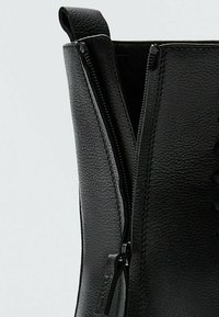 Gros plan d'une botte en cuir noir avec une fermeture éclair noire partiellement dézippée et une languette de tirage sur fond clair.