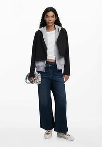 Musta ja harmaa lyhyt huppari, valkoinen paita, leveälahkeiset siniset farkut ja valkoiset kengät. Pitelee kuviollista käsilaukkua. Sileä materiaali.