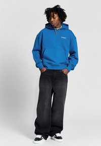 Blue cotton hoodie with front pocket and ribbed cuffs, paired with black wide-leg pants and black-and-white sneakers.