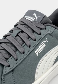 Sapatilhas cinza em camurça com o logotipo branco da Puma, atacadores texturizados e língua acolchoada. O sapato apresenta uma sola de borracha e um design elegante.