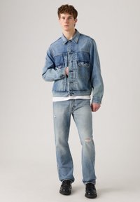 Young man wearing a light blue denim jacket and jeans with light distressing on a neutral background, standing with one hand partly inside the jacket.