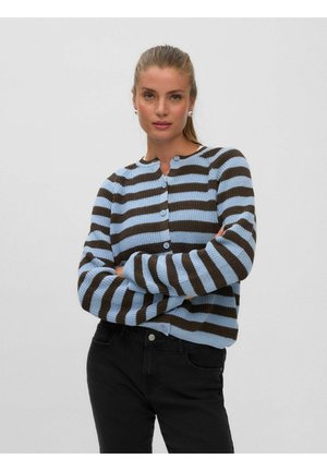 Donna con capelli castano chiaro raccolti in una coda di cavallo, indossa orecchini di perle e un maglione a righe blu e nero abbottonato con pantaloni neri, braccia incrociate.