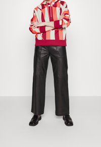 Felpa con strisce verticali rosse, corallo e crema; presenta una tasca frontale a marsupio e cappuccio con coulisse. Pantaloni a gamba larga in pelle nera. Scarpe nere.