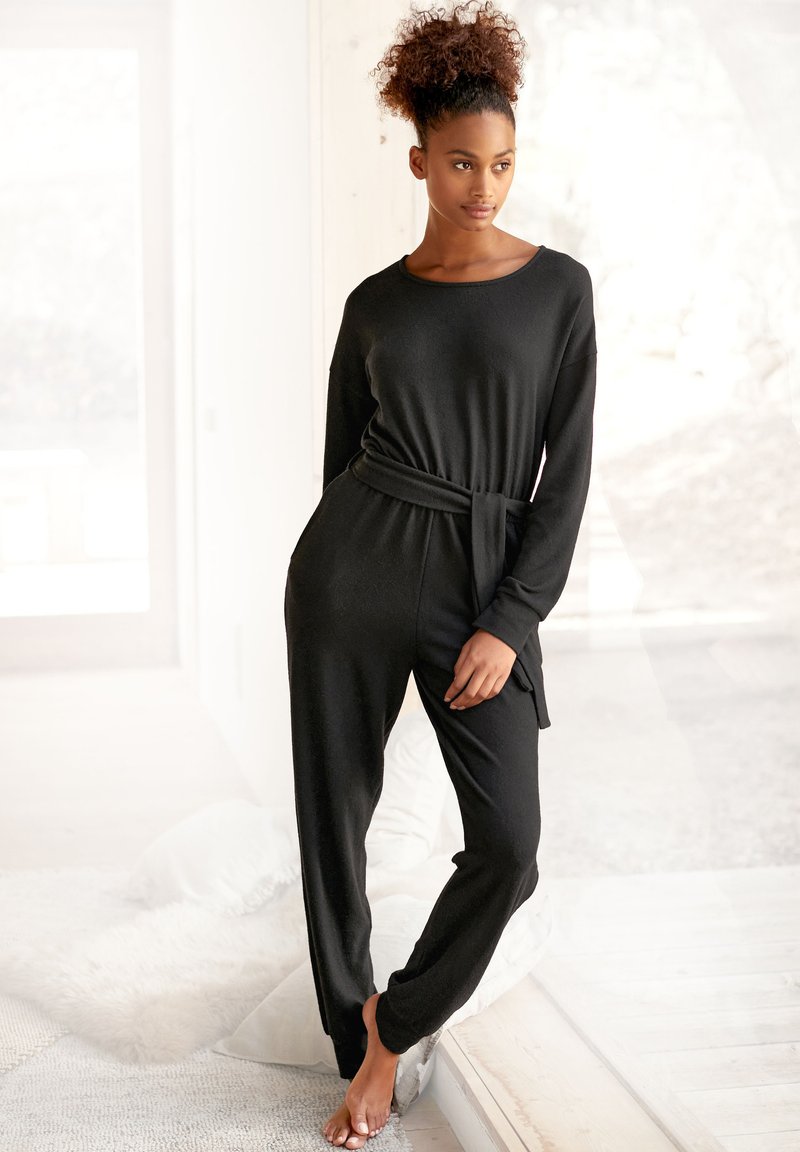 Zwarte, lange jumpsuit met een relaxte fit, strik taille-detail, zachte textuur en rechte pijpen, gedragen met blote voeten. Heldere, luchtige achtergrond.