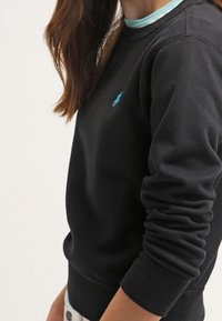 Svart sweatshirt i bomull med långa ärmar, som har en blå broderad logotyp på bröstet och en kontrasterande ljusblå krage.