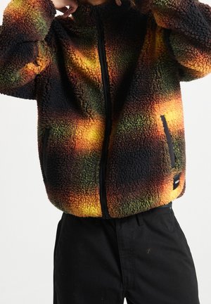 Person iført en flerfarvet sherpa fleecejakke med orange, gule og grønne nuancer og sorte bukser mod en ensfarvet baggrund.