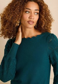 Bordo pulover s izrezom u obliku školjke i teksturiranim, uzorkovanim rukavima. Mekana pletenina s detaljnim vezom i uskim manžetama.