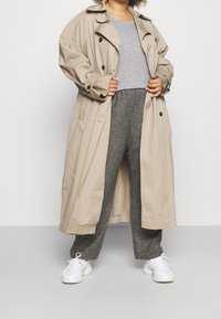 Manteau beige avec une taille ceinturée, grand col et fermetures à boutons, associé à un haut gris et un pantalon texturé gris foncé. Baskets blanches.