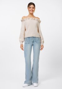 Off-the-shoulder beige sweater with ruffled edges, paired with light blue flared jeans and white sneakers. Smooth texture, casual fit.