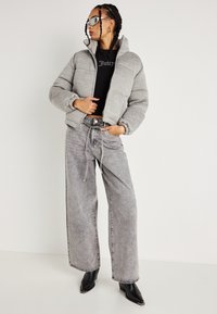 Kvinde med fletninger har sorte solbriller på, en grå puffjakke, en sort crop top, grå bukser med vide ben og sorte spidse støvler, står sidelæns.