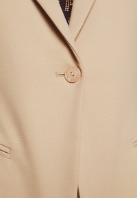 Blazer beige con un solo botón redondo abrochado, con solapas de muesca y bolsillos de ojal a cada lado.