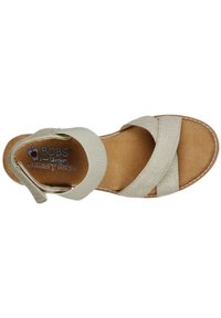 Beige sandaal met twee gekruiste stofbanden, een verstelbare achterband en een voetbed van traagschuim, met een gebogen zool en gestikte accenten.