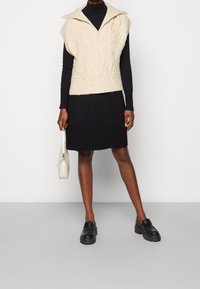 Maglione senza maniche a maglia cable color crema con un ampio colletto, abbinato a una gonna nera. La modella tiene in mano una borsa bianca testurizzata e indossa scarpe nere.