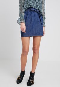 Jupe mini en denim bleu foncé avec taille ceinturée. Texture lisse, coupe droite et ourlet fini. Portée avec des bottines noires ornées d'accents dorés.