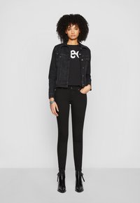 Chaqueta de mezclilla negra con acentos metálicos, combinada con una camiseta negra ajustada que presenta texto en blanco, y jeans negros ajustados. Botines negros completan el atuendo.