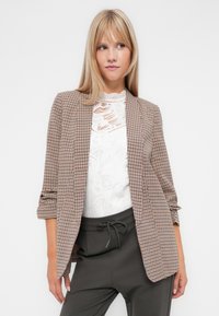 Le blazer présente un motif pied-de-poule marron et crème, une coupe ajustée, une ouverture à l'avant et des manches roulées, superposé à un haut en dentelle blanc.