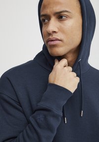 Sudadera azul marino con capucha, con puños acanalados, cordones ajustables y detalle de logotipo bordado en la manga. Textura suave.