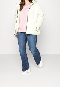 Pufa bunda světle zelené barvy, zapínaná na zip s kapucí, v kombinaci s růžovou košilí a modrými džíny. Bílé tenisky dotvářejí celý outfit.
