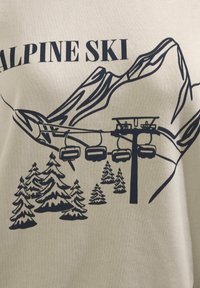 T-shirt in cotone beige con una grafica blu scuro di montagne, una seggiovia con tre posti e alberi di conifere stilizzati. Il testo recita "ALPINE SKI."