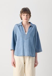Camicia in denim azzurro chiaro con scollo a V, maniche a tre quarti e orlo grezzo. Presenta dettagli di cucitura sottili sul colletto e sulle spalle.