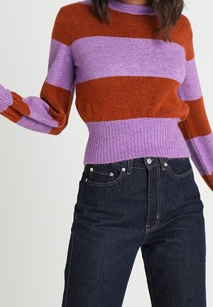 Personne portant un pull rayé avec de larges bandes violettes et orange rouille et un jean taille haute foncé sur un fond uni.