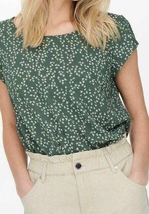 Frau trägt eine grüne Kurzarmbluse mit weißem Blumenmuster und eine beige Hose mit hohem Bund, Hände in den Taschen, Oberkörper sichtbar.