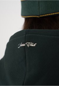 Mørkegrønn hettegenser med høy krage og brodert "Special Club"-logo i hvitt på baksiden. Teksturert stoff med synlig søm.