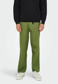 Pantalon en coton vert à coupe droite, avec un cordon de serrage à la taille. Porté avec un pull noir et des baskets blanches.