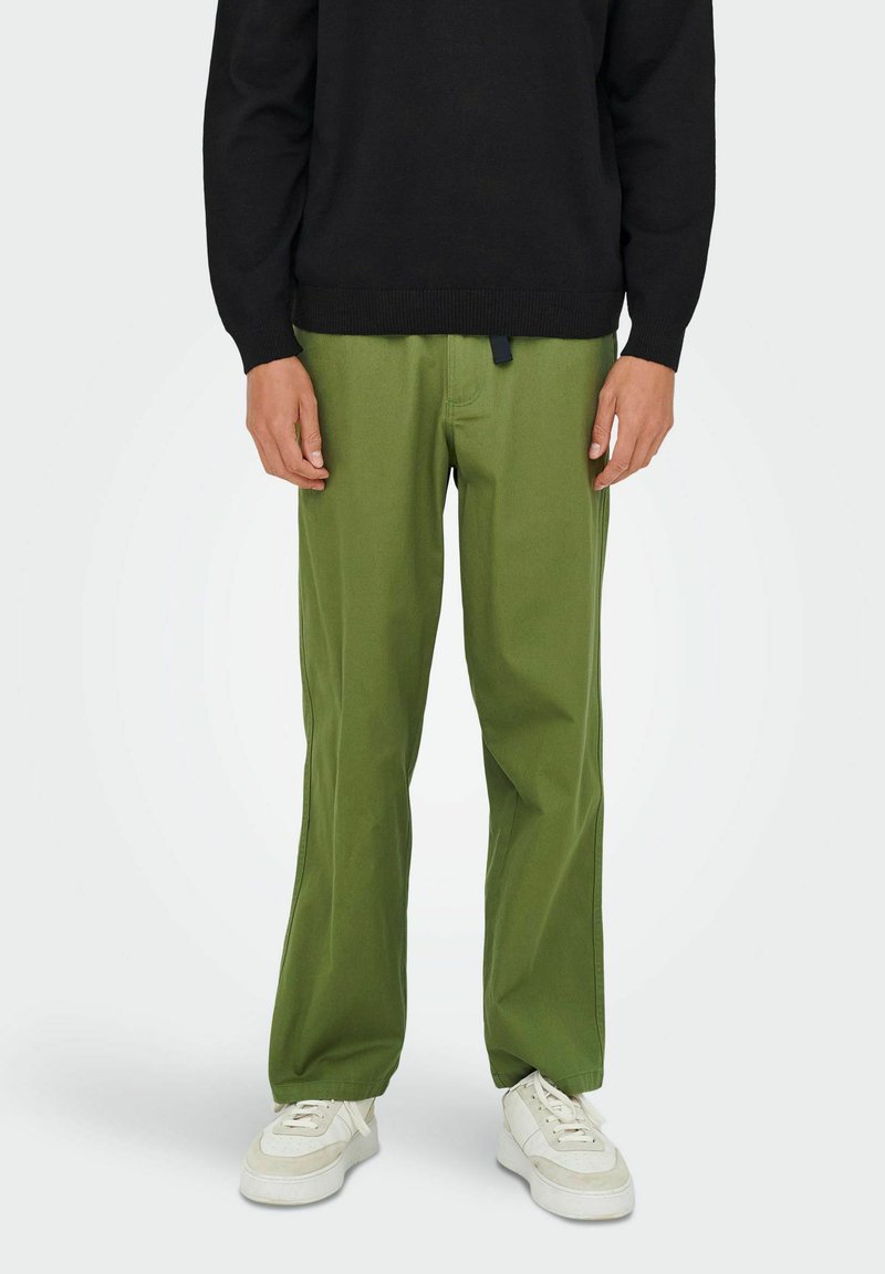 Pantalon en coton vert à coupe droite, avec un cordon de serrage à la taille. Porté avec un pull noir et des baskets blanches.