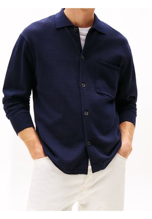 Mann trägt marineblauen, knöpfbaren Cardigan mit Brusttasche über weißem Hemd, Hände in den Taschen, kombiniert mit hellen Hosen.