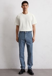 Biała koszulka z krótkim rękawem zestawiona z jasnoniebieskimi dżinsami i czarnymi butami na sznurówki, umieszczona na neutralnym tle.