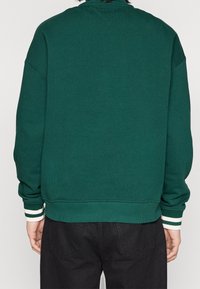 Sudadera verde con cuello y puños acanalados, con rayas blancas, y un corte relajado. Costuras visibles y una superficie texturizada.