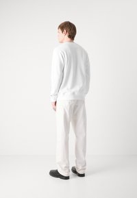 Sudadera blanca con puños y dobladillo acanalados, combinada con pantalones beige y zapatos negros, vista desde atrás sobre un fondo neutro.