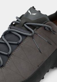 Pelēki āra apavi ar gludu un teksturētu materiālu kombināciju, izceļoties ar izteiktu diagonālo šuvju un atstarotājām auklām. Ūdensnecaurlaidīgs GORE-TEX.