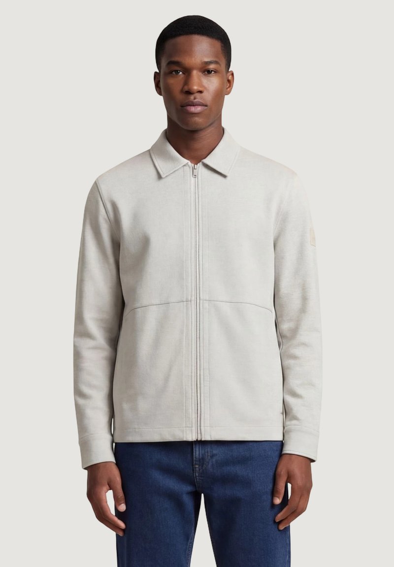 Jeune homme portant une veste zippée beige clair avec col et un jean bleu, debout devant un fond clair uni.