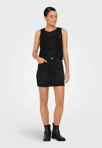 Zwart gehaakt vest met een vastgebonden detail aan de voorkant over een zwart top, gecombineerd met een nauwsluitende zwarte spijkerrok en zwarte enkelboots.