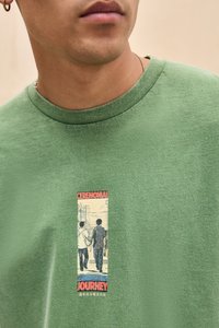 Man wearing green shirt with graphic showing two people holding hands, text "Ceremonial Journey" and small Japanese characters below.